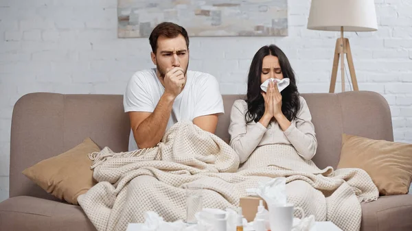 Sick woman sneezing while man couching near bottles with pills on coffee table - foto de stock