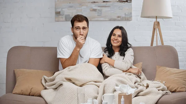 Brunette woman smiling near sick boyfriend coughing in living room — Stock Photo