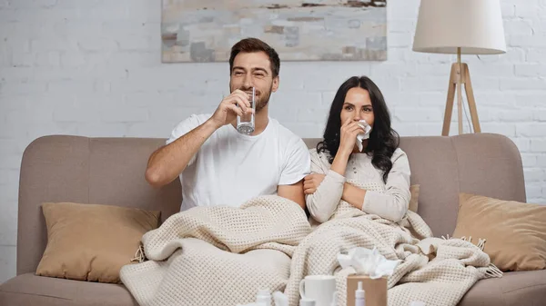 Sick woman with napkin near smiling man drinking water in living room - foto de stock