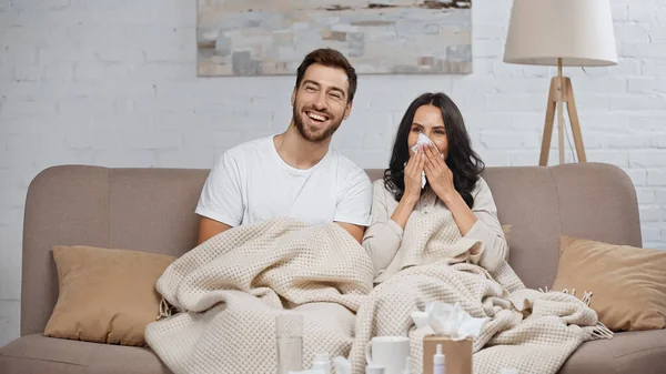 Bearded man smiling near sick girlfriend sneezing in tissue — Stock Photo