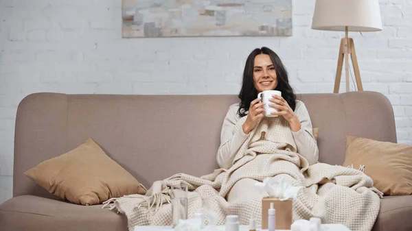 Kranke brünette Frau hält Tasse Tee und lächelt, während sie auf der Couch sitzt — Stockfoto