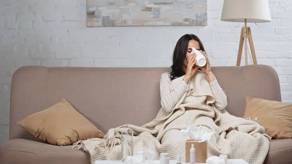 Kranke brünette Frau hält Tasse und trinkt Tee, während sie auf der Couch sitzt — Stockfoto