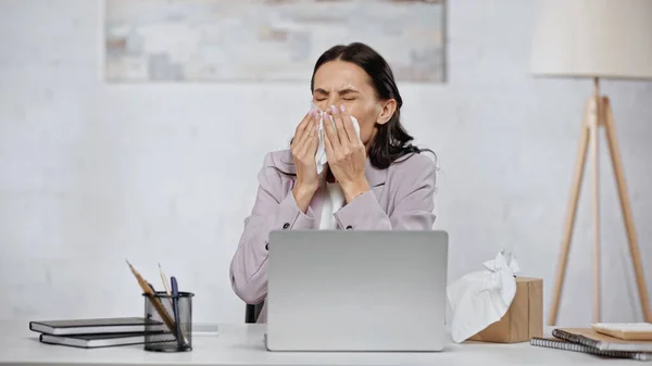 Allergic businesswoman sneezing in tissue near laptop on desk — Photo de stock