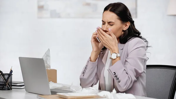 Brünette Geschäftsfrau mit Allergie niest, während sie auf dem Schreibtisch neben dem Laptop auf dem Smartphone spricht — Stockfoto