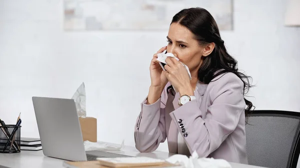 Brünette Geschäftsfrau mit Allergie niest in Serviette, während sie in der Nähe des Laptops auf dem Schreibtisch mit dem Handy spricht — Stockfoto