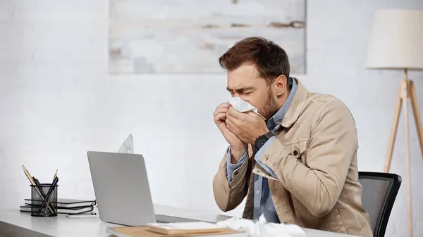 Аллергический бизнесмен чихает в салфетке рядом с ноутбуком на столе — стоковое фото