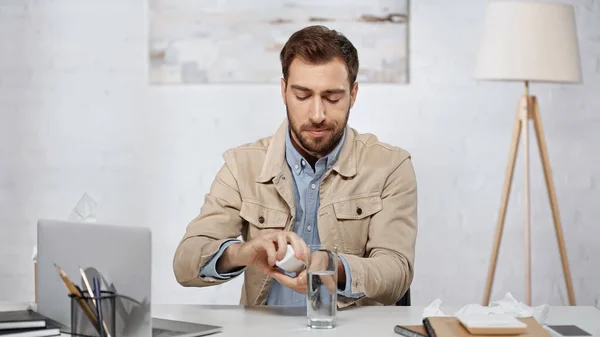 Allergic businessman holding bottle with pills near laptop on desk - foto de stock