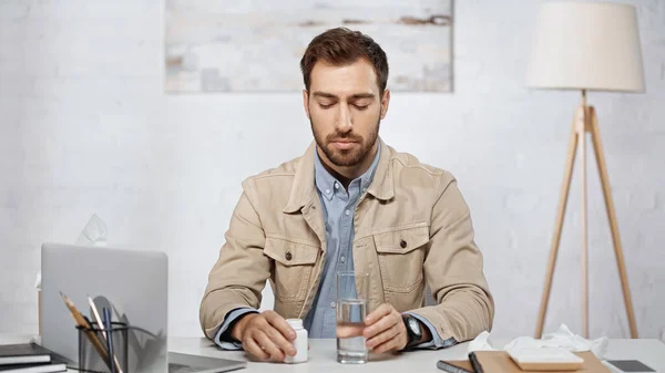 Allergic businessman looking at bottle with pills near glass of water and laptop on desk - foto de stock