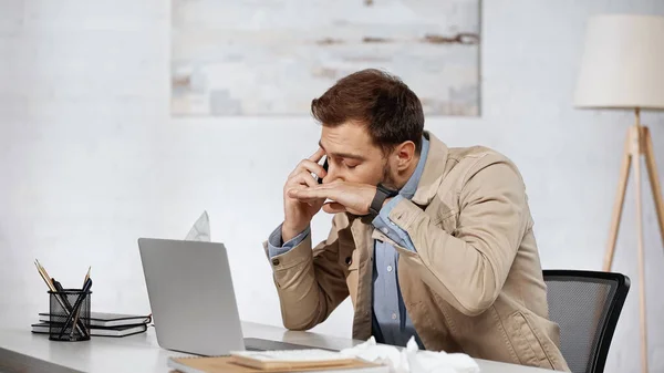 Allergic businessman sneezing while talking on smartphone near laptop on desk — Photo de stock