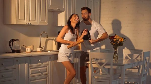 Happy man with frying pan dancing near joyful girlfriend with paper mill — Fotografia de Stock