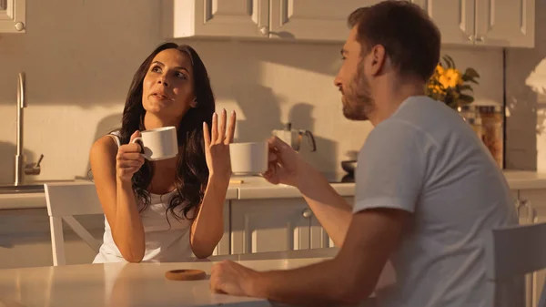 Nachdenkliche junge Frau hält Tasse Kaffee in der Hand und spricht mit verschwommenem Freund — Stockfoto
