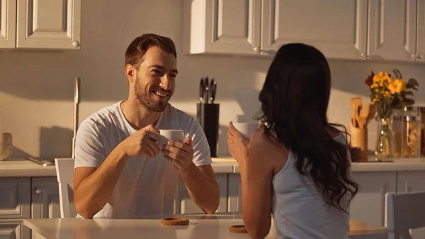 Glücklicher Mann mit Tasse und Blick auf brünette Freundin in der Küche — Stockfoto