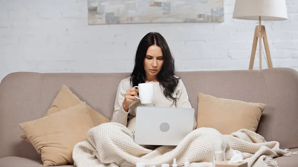 Kranke Frau sitzt mit Laptop und hält Tasse Tee — Stockfoto