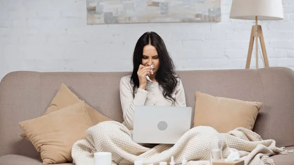 Sick young woman sneezing in tissue while looking at laptop — Stock Photo