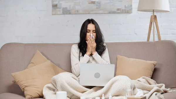 Ill woman sneezing in tissue while looking at laptop — Stock Photo