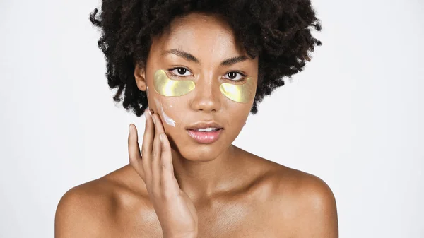 Femme afro-américaine avec des patchs pour les yeux appliquant de la crème sur le visage isolé sur gris — Photo de stock