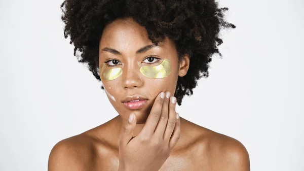 Junge afrikanisch-amerikanische Frau mit goldenen Augenflecken, die kosmetische Creme isoliert auf grau aufträgt — Stockfoto