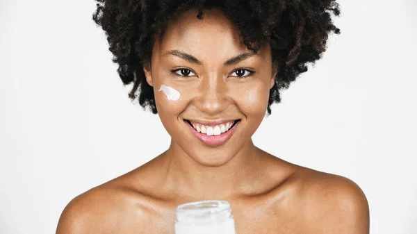 Sorrindo mulher americana africana com creme no rosto olhando para a câmera perto frasco borrado isolado em cinza — Fotografia de Stock