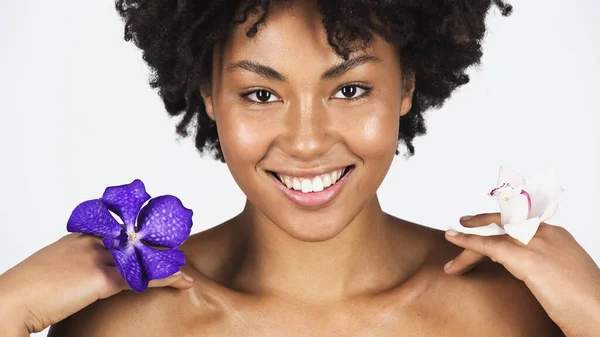 Pretty african american woman smiling while holding orchid flowers isolated on grey — Stock Photo