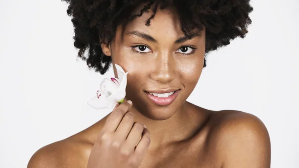 Smiling african american woman holding orchid near cheek isolated on grey — Stock Photo