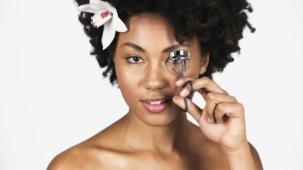 Young african american woman with orchid in hair using eyelash curler isolated on grey — Stock Photo