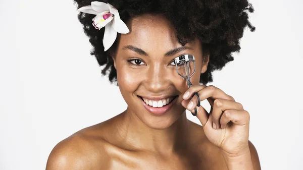 Smiling african american woman with flower in hair using eyelash curler isolated on grey — Stock Photo