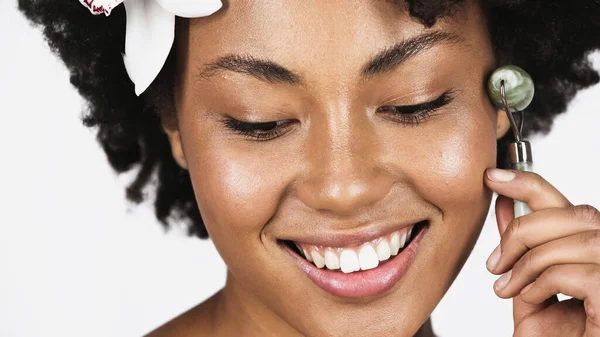 Vue rapprochée de la femme afro-américaine souriante avec fleur dans les cheveux à l'aide d'un rouleau de jade isolé sur gris — Photo de stock