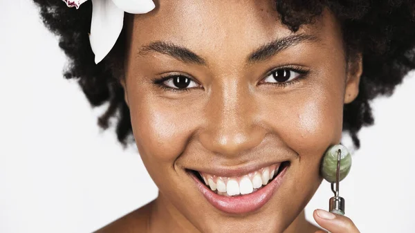 Close up view of smiling african american woman with flower in hair using jade roller isolated on grey — Stock Photo