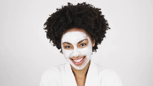 Mulher afro-americana positiva em roupão de banho branco e máscara facial olhando para a câmera isolada em cinza — Fotografia de Stock