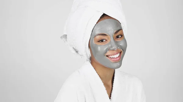 Africano mulher americana com máscara de barro no rosto e toalha na cabeça sorrindo isolado no cinza — Fotografia de Stock