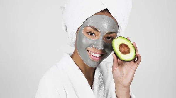 Mujer afroamericana sonriente con máscara de arcilla en la cara sosteniendo aguacate aislado en gris - foto de stock