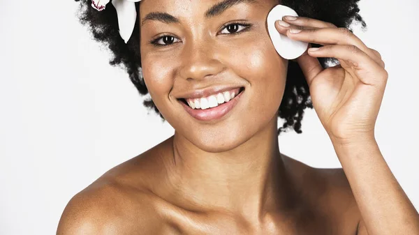 Afroamerikanerin mit Orchidee im Gesicht zur Haarreinigung mit Wattepad isoliert auf grau — Stockfoto
