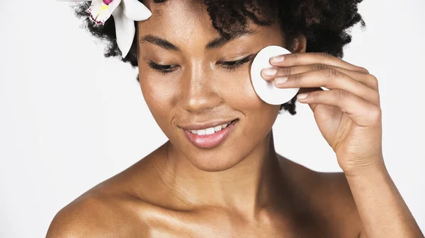Jeune femme afro-américaine avec orchidée dans la joue de nettoyage des cheveux avec coton isolé sur gris — Photo de stock