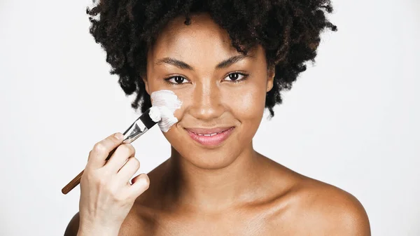 Жіноча рука наносить маску для обличчя на усміхнену афроамериканку ізольовану на сірому — стокове фото