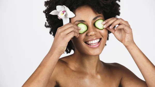 Fröhliche afrikanisch-amerikanische Frau mit Orchideenblume im Haar, die Gurkenscheiben in Augennähe hält, isoliert auf grau — Stockfoto