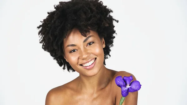 Mujer afroamericana sonriente con piel limpia sosteniendo orquídea aislada en gris - foto de stock