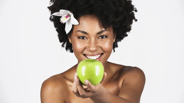 Positive african american woman with orchid in hair holding apple on blurred foreground isolated on grey — Stock Photo