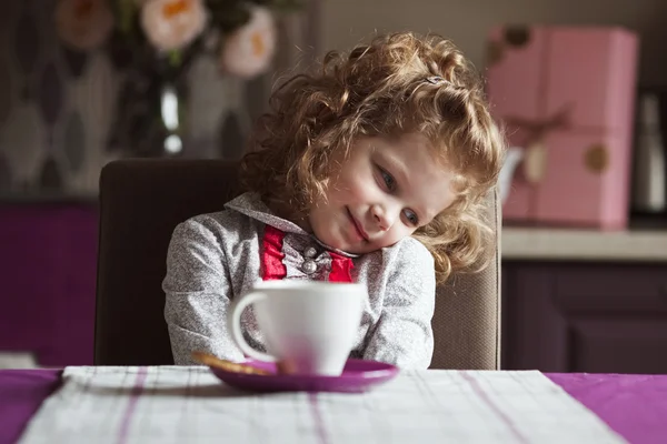 Pequena menina encaracolado com copo sentado à mesa — Fotografia de Stock