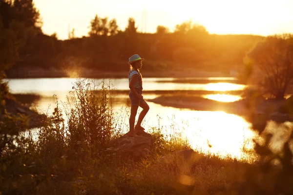 Dziewczyna w kapeluszu stoi na brzegu rzeki na zachodzie słońca — Zdjęcie stockowe