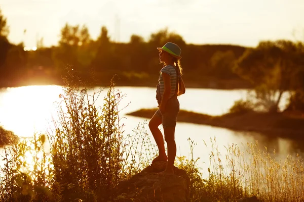 Little girl in hat stands on the river bank at sunset — Stock Photo, Image