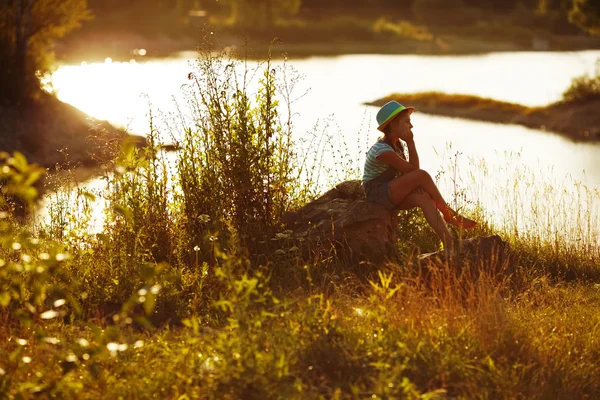 Girl in hat sitting on the bank of the river — Stock Photo, Image