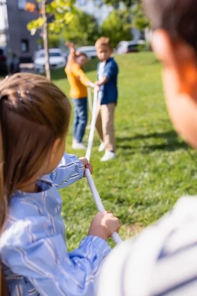 Rope in hands of children playing tug of war on blurred background in park