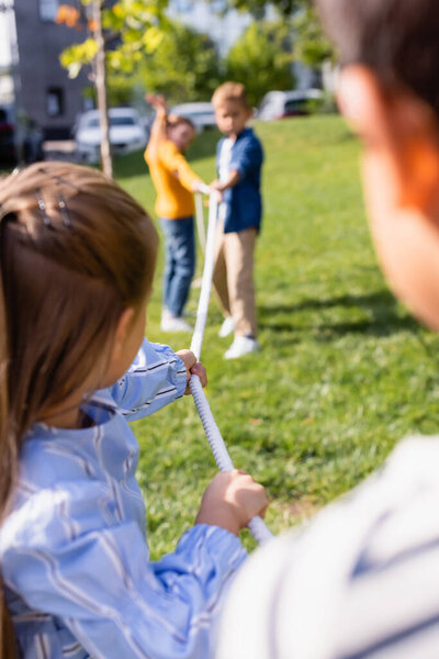 Rope in hands of children playing tug of war on blurred background in park 