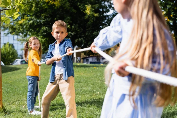 Boy pulling rope while playing tug of war with friends on blurred foreground in park