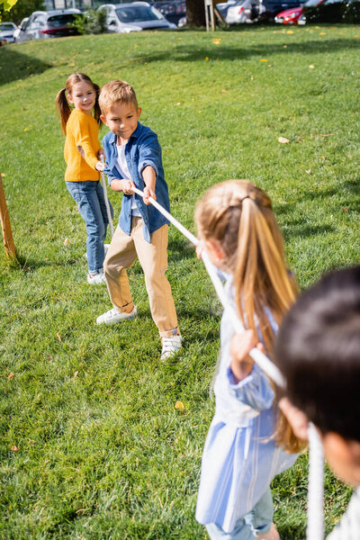 Cheerful kids plying tug of war with friends on blurred foreground in park 