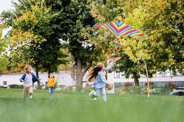 Girl looking at flying kite while running near friends on blurred background on lawn 