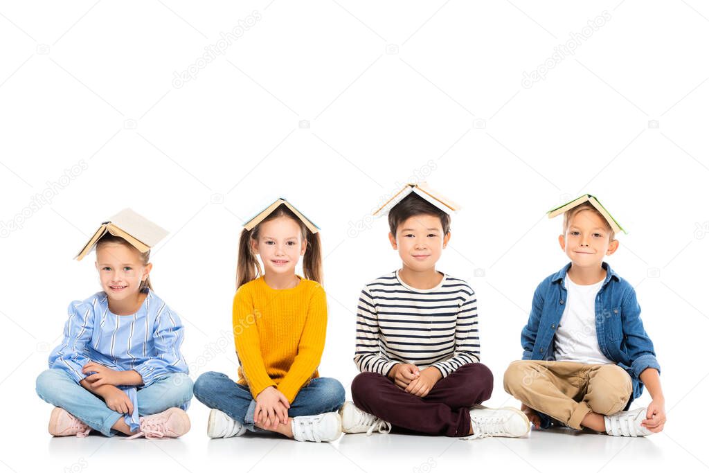 Smiling multiethnic kids with books on heads sitting on white background 