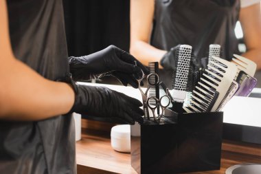 Plastik eldivenli kuaförün kısmi görüntüsü, arka planda bulanık taraklar olan makas tutacağı takıyor.