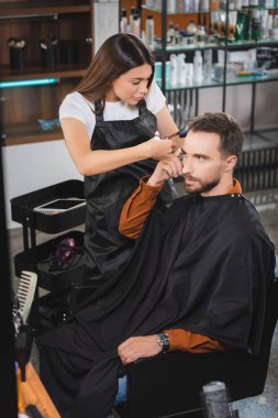 Önlüklü genç kuaför parmağıyla işaret eden adamın saçını kesiyor.