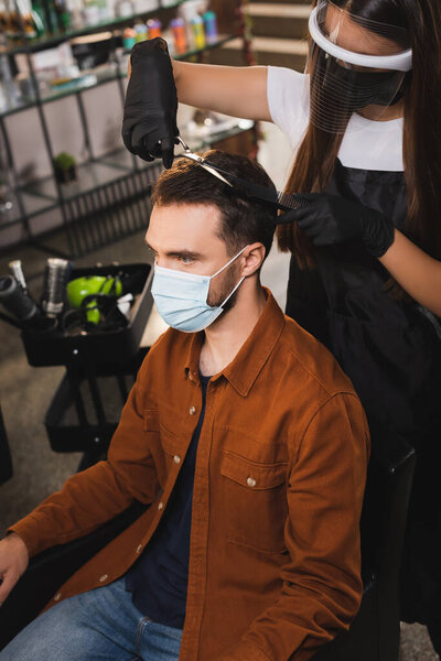 barber in face shield and latex gloves cutting hair of man in medical mask